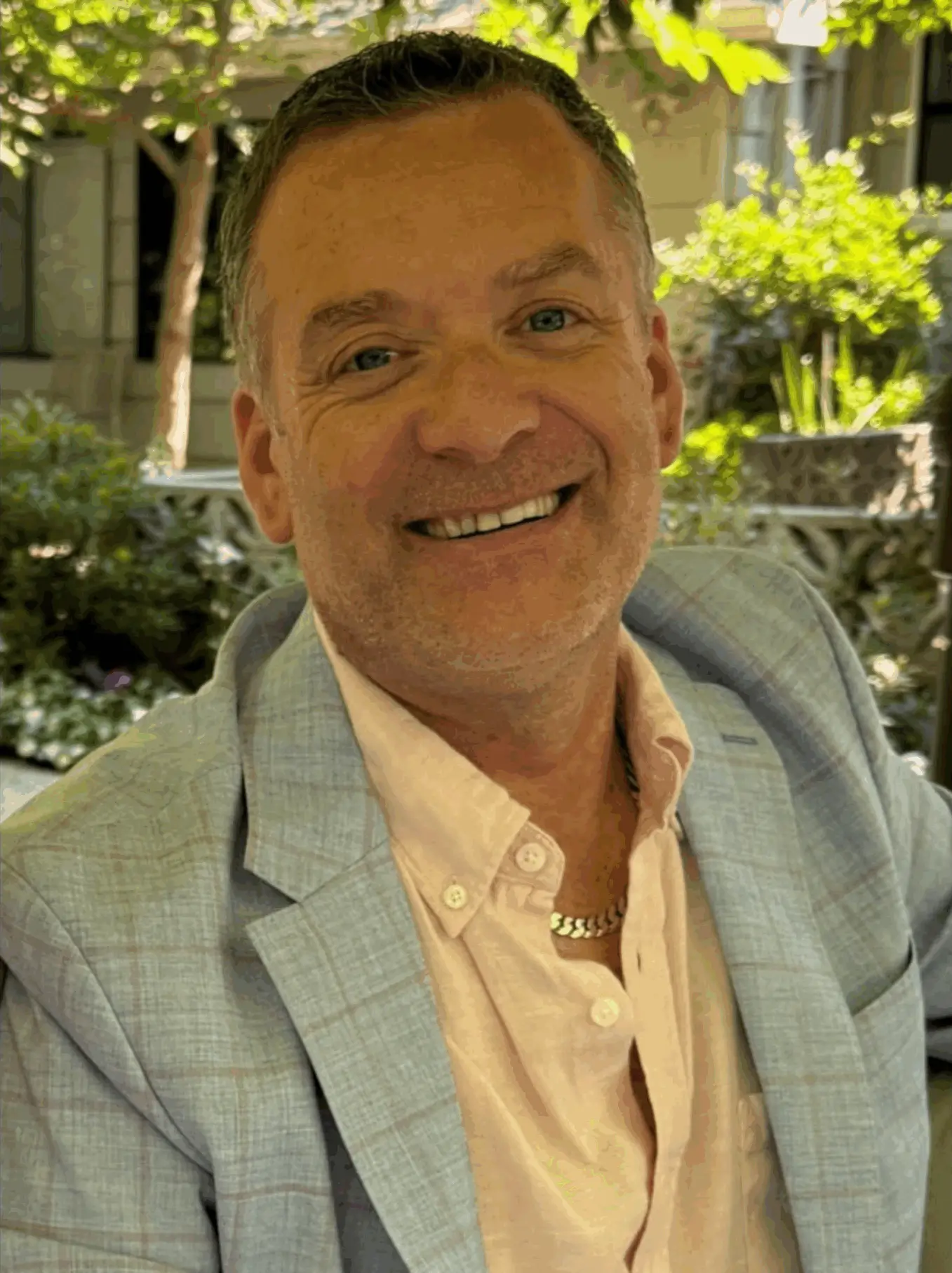 A smiling man in a light gray plaid blazer and peach shirt outdoors.